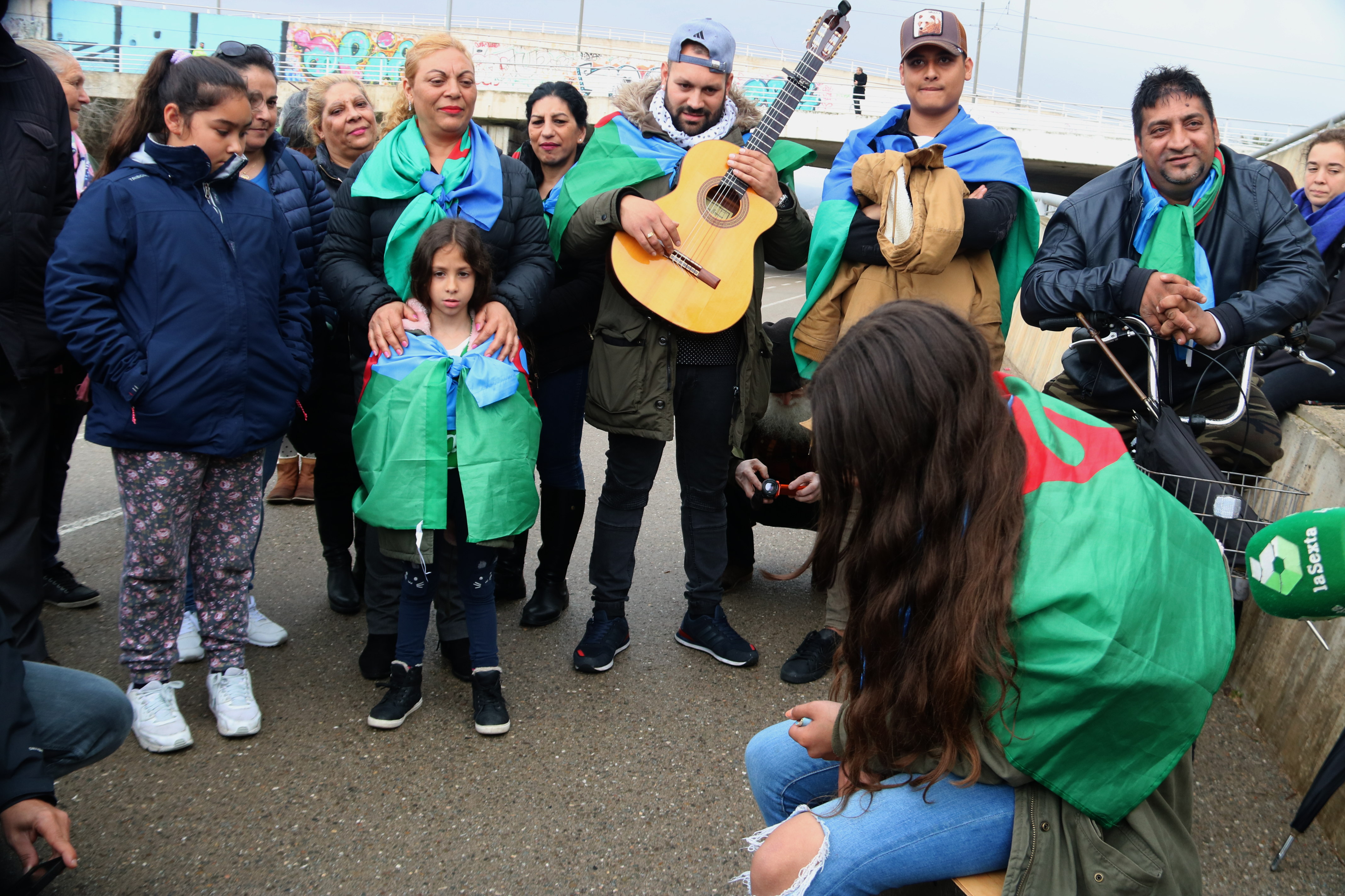 A girl, sitting on a 'cajón' sings 'Gelem, Gelem', the anthem of the Roma people, under the watchful gaze of a group of people on the banks of the Besòs River.