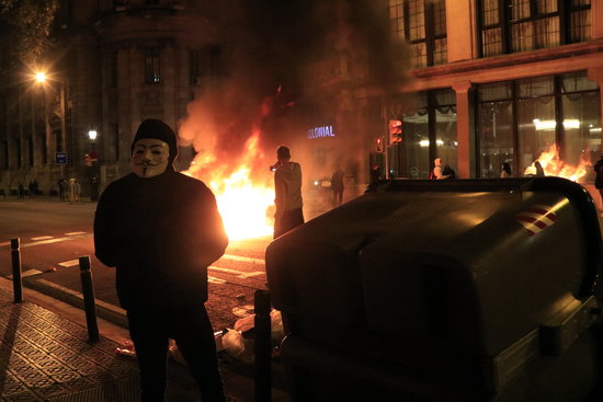 A trash can being burned down as a protesters wears an Anonymous group mask, on October 30, 2020 (by Laura Fíguls)