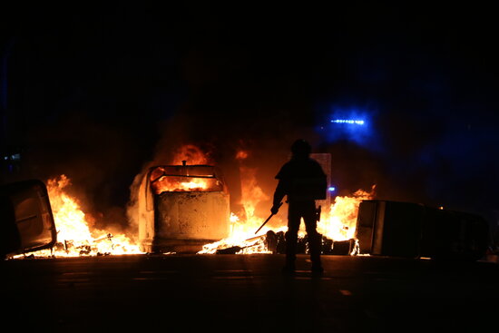 Silhouette of a Catalan riot police officer in front of several burnt-out containers during protests in Barcelona over the imprisonment of rapper Pablo Hasél, February 16, 2021 (by Miquel Codolar)