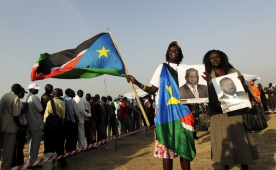 Two women showing photos of South Sudan leaders in front of a people waiting to vote (by Reuters)
