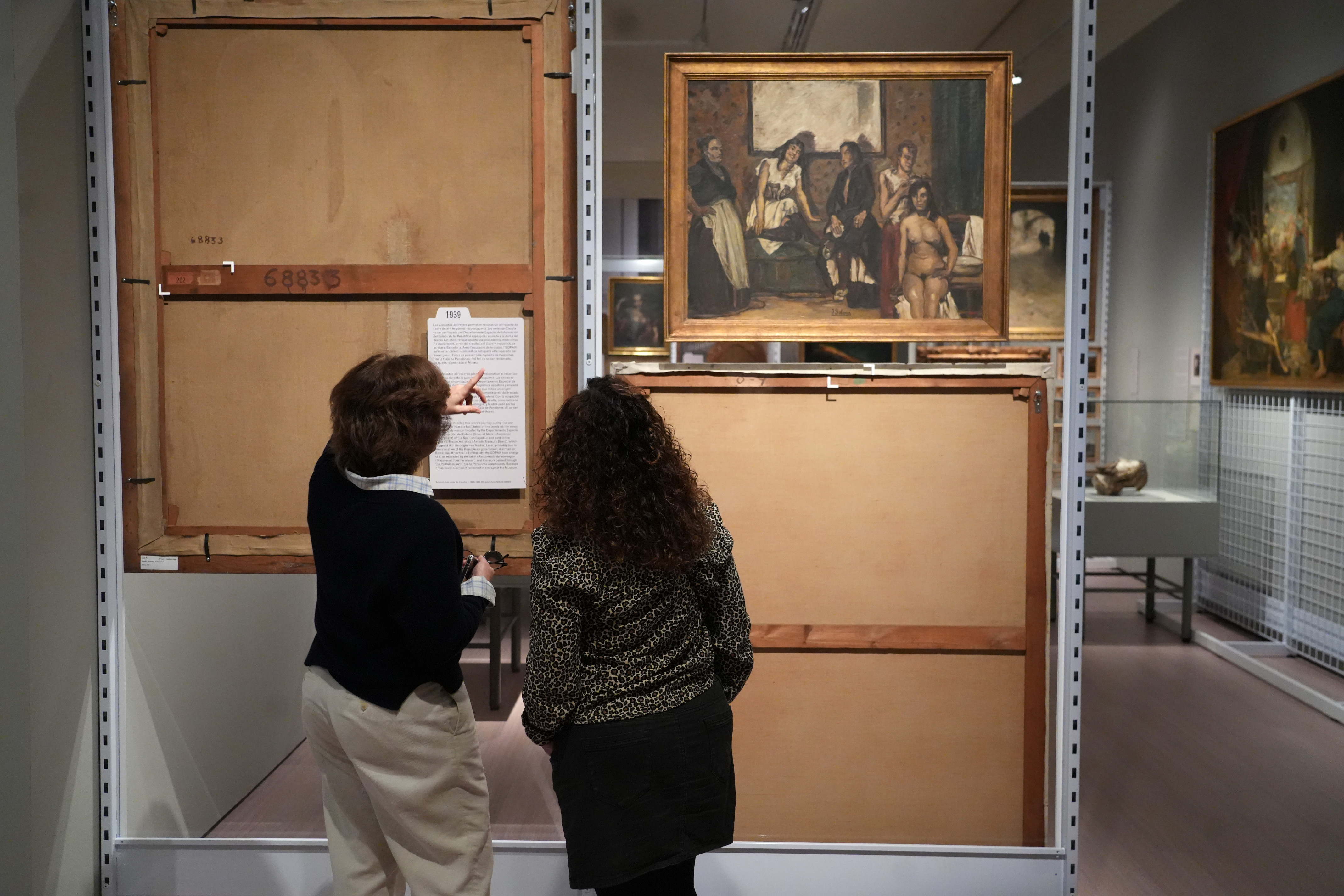Two women look at the labels on the back of a painting in the exhibition 'Recovered from the Enemy. The Francoist Deposits at the MNAC'