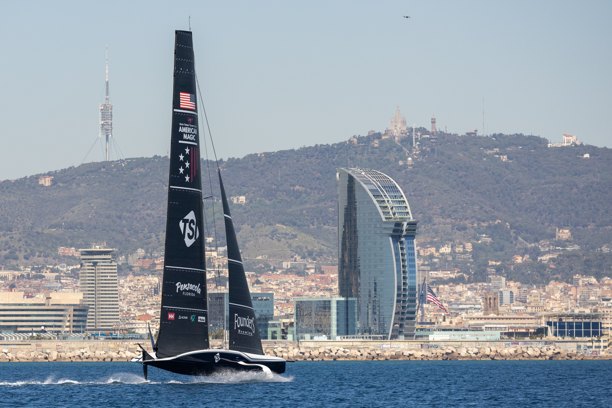 The AC75 of American Magic ahead of the America's Cup sail championship sails in front of Barcelona with the US flag waving