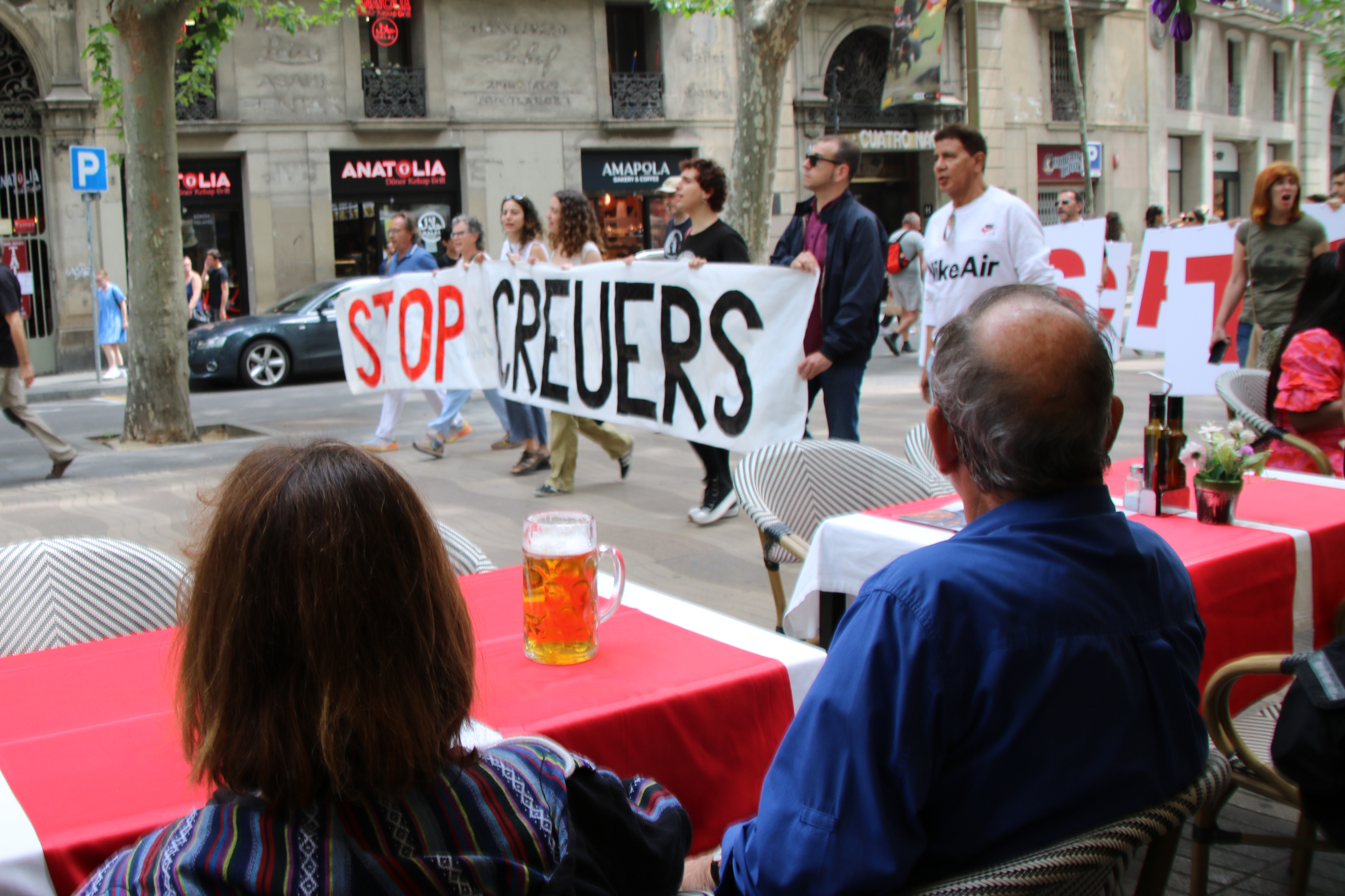 Two tourists enjoy a beer at a bar in La Rambla boulevard while observing residents demonstrate against cruises on May 7, 2023