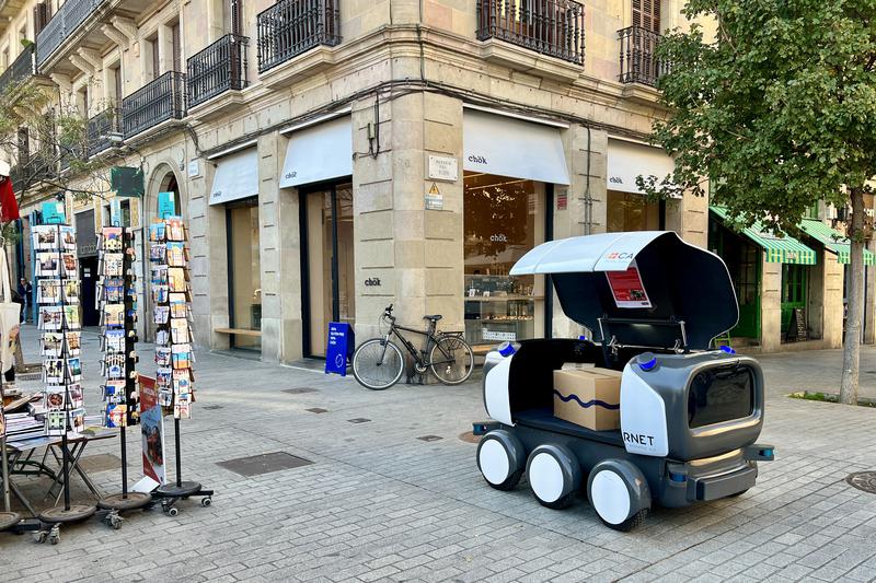 Robot Ona with a parcel inside next to a kiosk and a store in Barcelona during testing on November 19, 2024