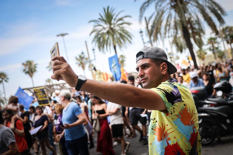 A tourist takes a selfie with an anti–mass tourism rally marching behind him in Barcelona, July 2024