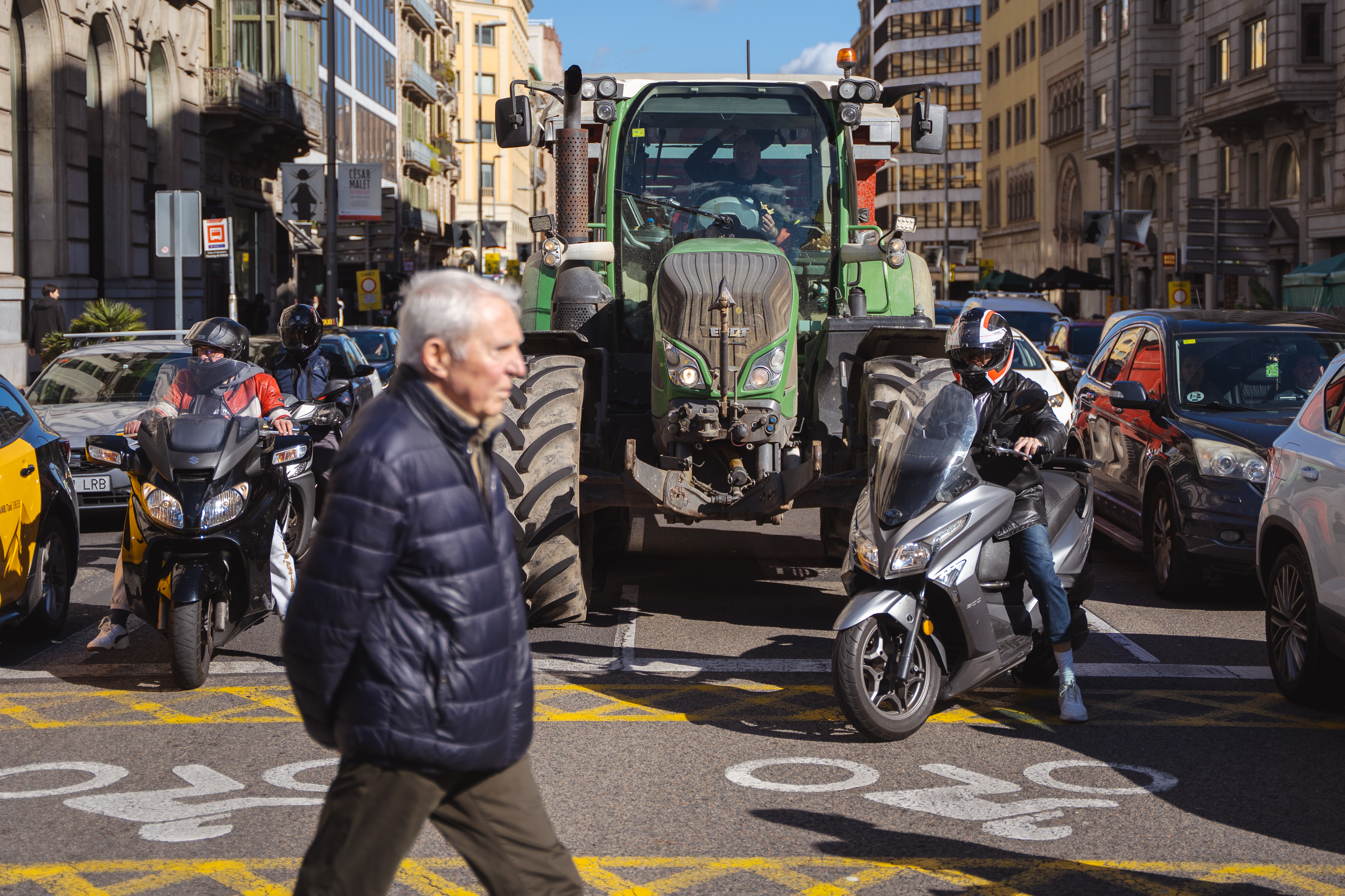 A man crosses the street as taxis, motorcycles, cars, and tractors wait behind him