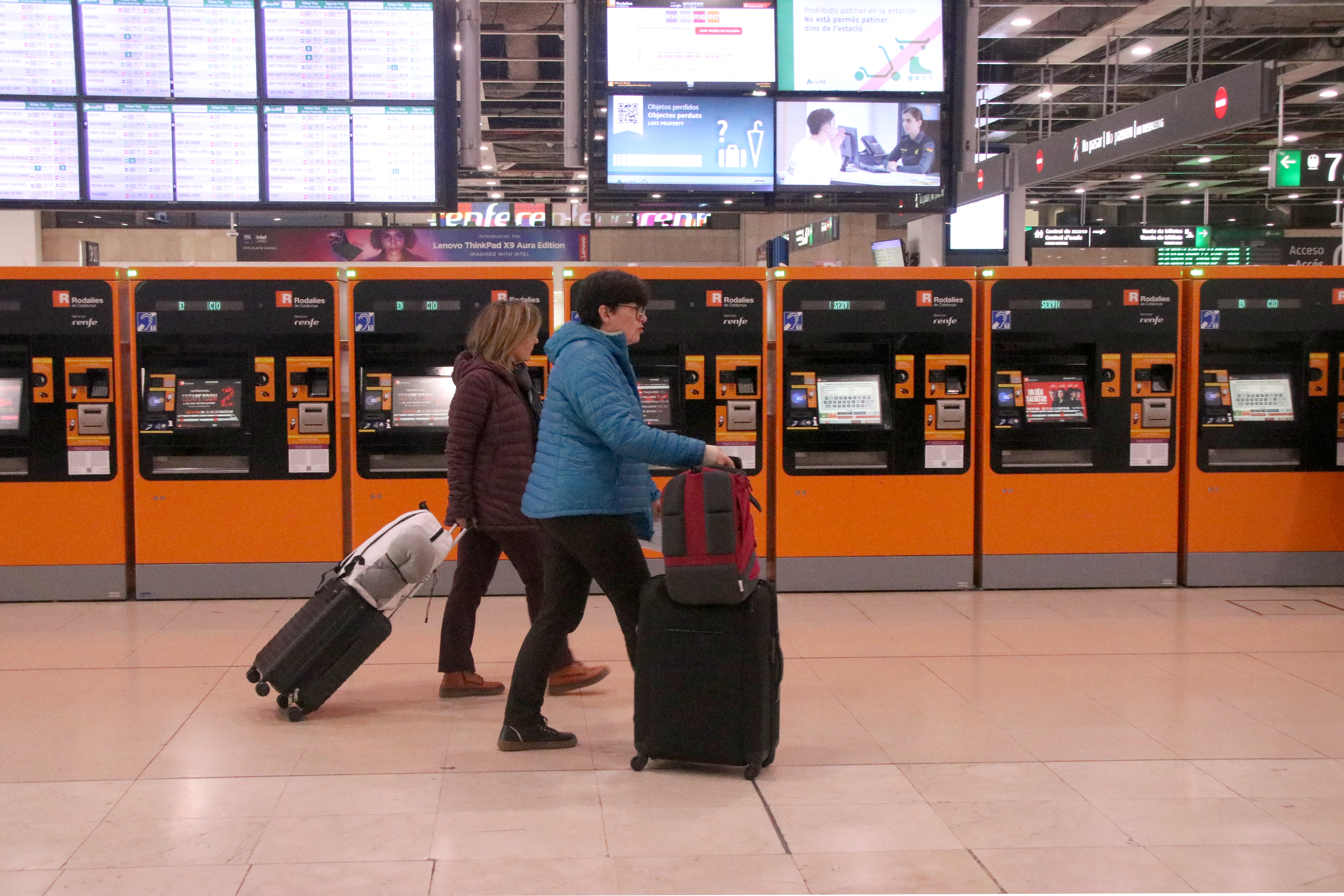 Two people with luggage walking around the Barcelona Sants train station on March 17, 2025