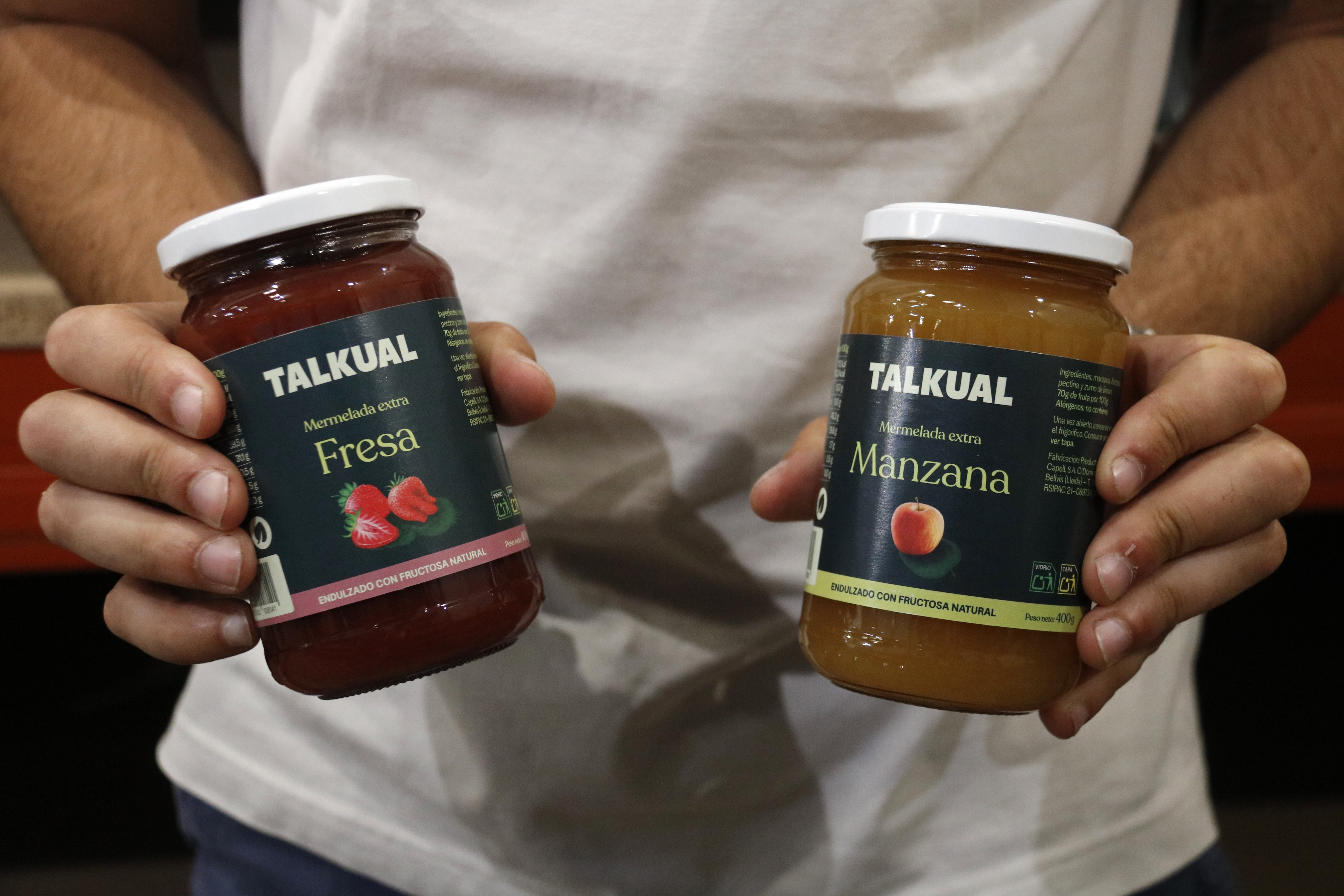 Two hands holding two jars of jam — one strawberry and the other apple — from the company Talkual