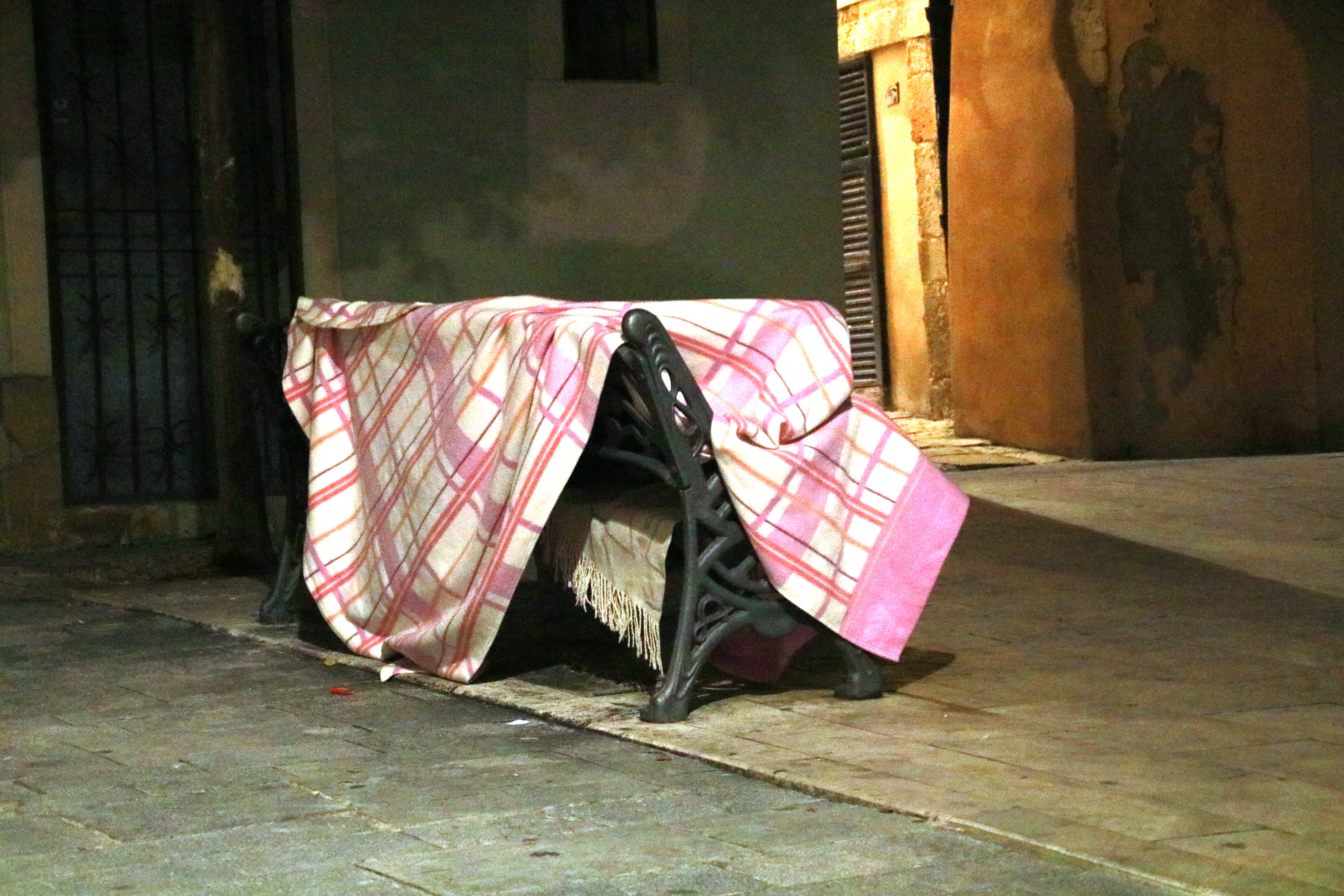 A homeless person covered with a blanket on a bench in a square in Tarragona
