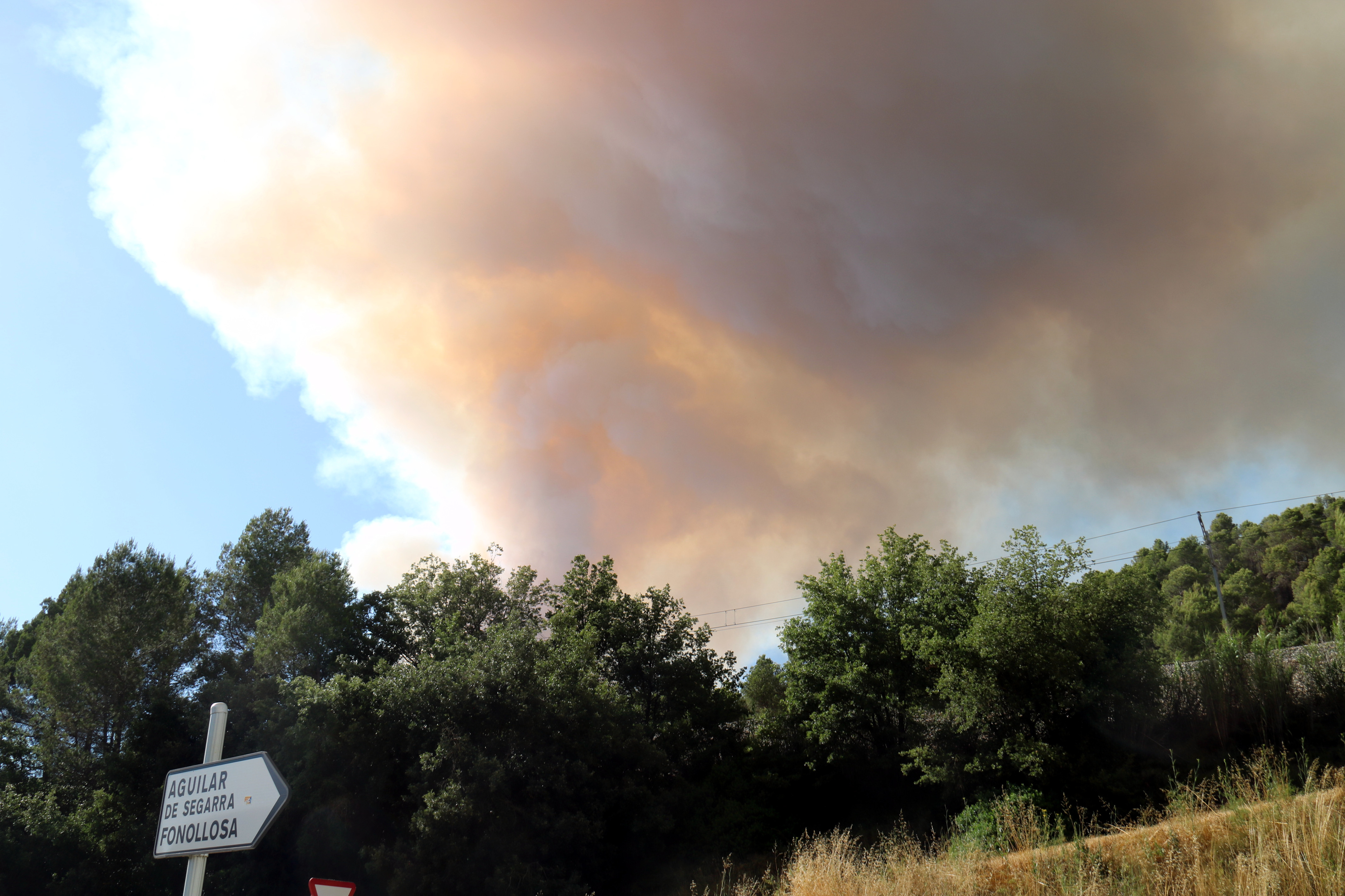 A large cloud of smoke from a wildfire in Sant Pere Sallavinera rises above a road sign pointing toward Aguilar de Segarra and Fonollosa